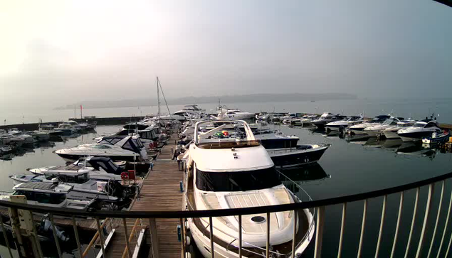 A view of a marina filled with various boats and yachts moored in calm water. The scene is hazy, with light fog covering the area. In the foreground, a large white motorboat is docked, with a wooden walkway leading to it. Several smaller boats are visible around it, all reflecting in the still water. The distant shoreline is faintly visible in the background, suggesting a tranquil setting.