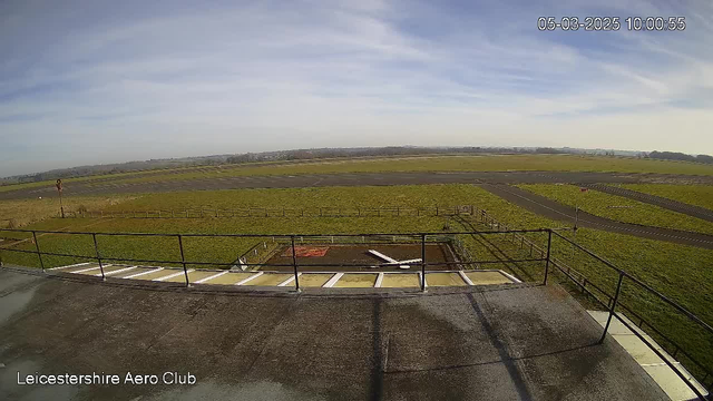 A view from a webcam at Leicestershire Aero Club, showing a wide, open grassy area with a runway on the left. In the foreground, there is a rooftop railing with steps leading down. The sky is mostly clear with some wispy clouds, and the landscape appears flat, extending toward the horizon. A few scattered trees are visible in the distance. The date and time in the corner indicate it is 10:00 AM on March 5, 2025.