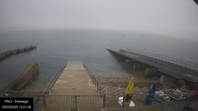 A foggy seascape is depicted, showing a partially visible, calm water surface. In the foreground, there are concrete steps leading down to the water, bordered by a metal railing. To the left, a small wooden pier extends into the water. Various boats in blue and yellow are positioned on the shore, alongside rocky terrain. The visibility is low due to fog, obscuring the horizon line and distant features.