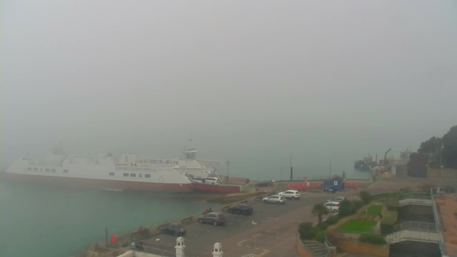 A foggy waterfront scene shows a large ferry boat docked at a pier. The visibility is low due to thick fog, obscuring much of the background. In the foreground, there are several parked cars in a lot, with a few scattered items on the ground. There are some green patches of grass and landscaped areas along the side of the parking lot. The overall atmosphere is quiet and misty.