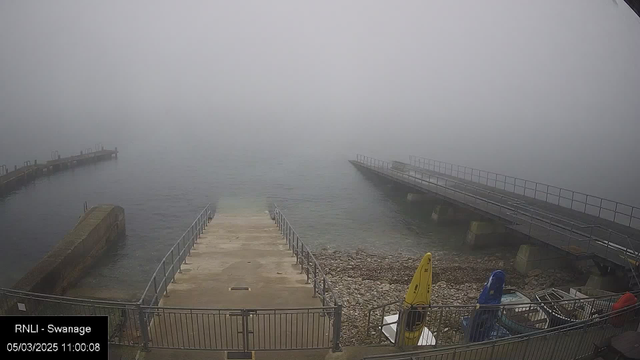 A foggy scene at a waterfront, with a partially visible wooden ramp leading down to the water. To the sides, there are two distinctive piers extending into the water. One pier has a railing, while the other appears to be stone. In the foreground, there are several boats, including a bright yellow kayak, resting on the shore near a rocky area. The overall visibility is low due to the dense fog, obscuring the horizon. The image is time-stamped at 11:00 on March 5, 2025.