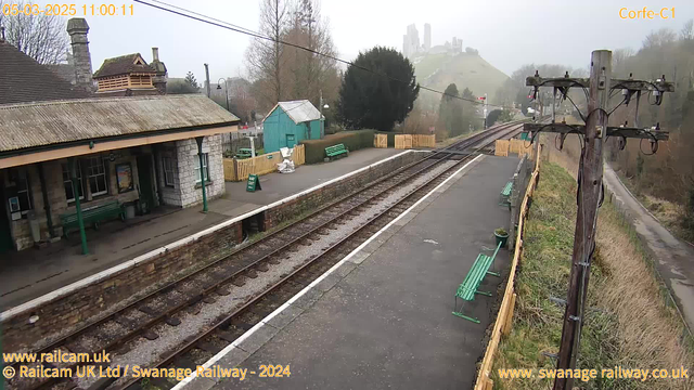 A foggy scene at Corfe Castle railway station. In the foreground, there are railway tracks next to a wide, paved platform lined with green benches. A stone building with a sloped roof is visible, featuring wooden beams and a large window. To the left, a small green shed and a wooden fence can be seen. In the background, the silhouette of Corfe Castle is visible atop a hill, partially obscured by mist, surrounded by trees and shrubbery.