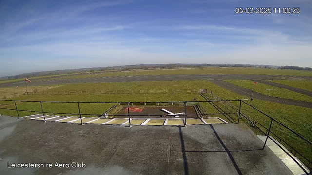 A view from the top of a structure overlooking an airfield. In the foreground, there is a railing and a flat, concrete surface. The airfield is mostly grassy with a few paved runways. A windsock is visible, indicating wind direction. In the background, the horizon features distant trees and hills under a clear blue sky with wispy clouds. The time is shown as 11:00 on March 5, 2025.