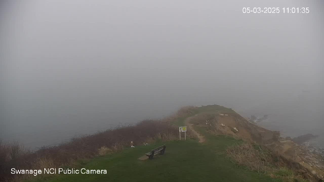 A foggy coastal view shows a grassy area with a path leading towards the edge of a cliff. A bench is positioned on the left side of the image, and there is a warning sign near the path. The background is obscured by thick fog, making it difficult to see the sea or any distant landscape. The overall atmosphere is misty and gray.