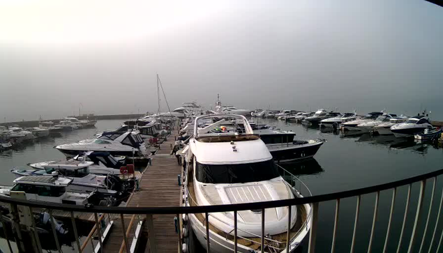 A foggy marina scene with a dock filled with various boats and yachts. In the foreground, a large white yacht with a shiny, polished surface is moored. The water is calm and reflects the shapes of the surrounding vessels. The background is obscured by fog, making it difficult to see distant boats and elements of the marina. The overall atmosphere is serene but slightly mysterious due to the low visibility.