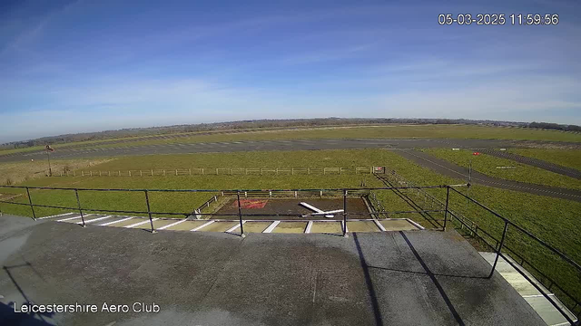 A clear blue sky is visible above a wide open area of grass and asphalt, indicating an airport or airfield. In the foreground, there is a railing from a building, with a concrete surface beneath it. Various flat areas of grass and sections of a tarmac runway can be seen, along with a fence bordering the grass. A small red object is present on the runway in the distance. The scene suggests a tranquil day at an aeroclub, with no clouds in the sky. The time is shown as 11:59:56 on March 5, 2025.