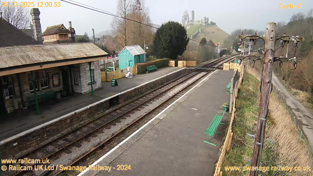 A railway station platform is depicted, featuring a dark stone building with a sloping roof on the left. Green benches are placed on the platform. A light blue shed is positioned towards the center, surrounded by wooden fencing. The railway tracks stretch into the distance, leading towards a hill with ruins visible at the top. There are utility poles and signals along the track. The scene is set on a clear day with limited visibility of the surrounding landscape.