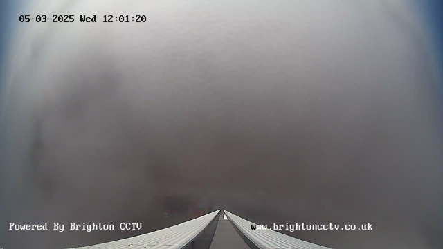 A view from a webcam showing a long white structure leading towards a foggy, grey horizon. The time displayed is 12:01:20 on March 5th, 2025. The text at the bottom indicates that the image is powered by Brighton CCTV, with a website link included. The scene is mostly filled with a uniform grey cloud cover, providing no distinct features in the background.