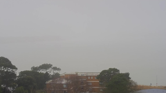 A foggy scene with low visibility over a body of water. In the foreground, there are some buildings with a reddish-brown exterior, partially obscured by trees and fog. The outlines of distant land or structures are barely visible in the haze, creating a muted, gray atmosphere. The natural landscape is blurred, suggesting a sense of stillness and quietness.