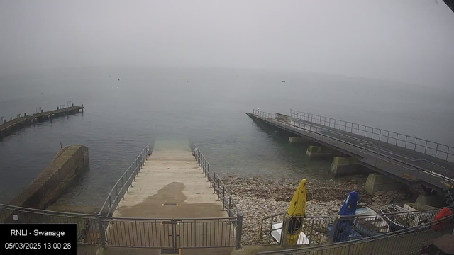 A foggy scene at a seaside location, showing a calm body of water with several small boats visible in the distance. In the foreground, there are two piers extending into the water on the left and right sides, with a ramp leading down to a rocky shore. A few kayaks in bright colors—yellow and blue—are parked near the edge. The atmosphere is overcast and hazy.