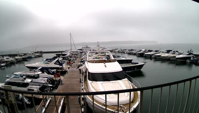 A marina filled with various boats and yachts is visible. The scene is overcast with a gray sky and fog, creating a misty atmosphere. A wooden dock is lined with boats of varying sizes, some parked side by side. In the background, further out on the water, more boats are seen, and land is faintly visible, suggesting a shore or coastline. The overall mood is calm and tranquil, with water gently reflecting the cloudy sky.