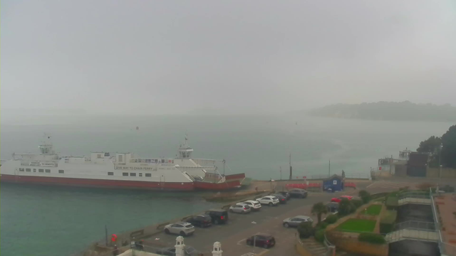 A cloudy, foggy scene depicting a ferry docked at a harbor. The ferry has a white body with a red bottom and visible markings on its side. The water is a muted blue-green tone, with gentle waves and some indistinct shapes in the distance, likely more boats or landforms. There are several parked cars in a lot beside the ferry, which features a few plants and landscaping elements in the foreground. In the background, there is a line of trees and a faint outline of land, blending into the mist.