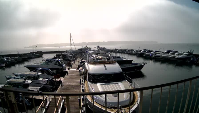 A marina filled with various boats docked in calm water. In the foreground, a large white and brown luxury yacht is prominent, with its roof visible. Surrounding it are smaller boats of different sizes and colors, including blue and white ones. The scene is set against a backdrop of foggy weather, with a blurred shoreline in the distance. The overall mood is serene and quiet, with muted lighting due to the fog.