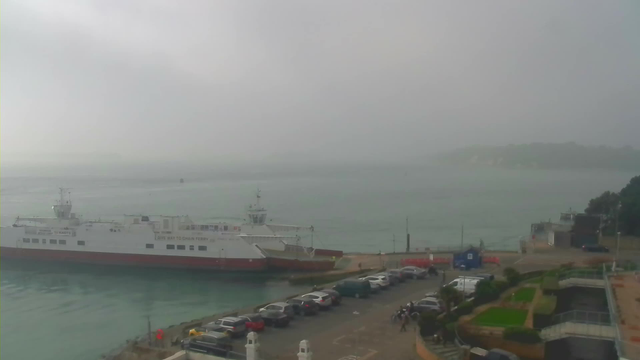 A foggy seaside view featuring a large ferry docked at a terminal. The ferry has a white body with a red bottom. The water is calm, and the background shows a muted landscape with trees and land partially obscured by fog. In the foreground, there is a parking area with several cars parked along the edge, and some people are walking nearby. The atmosphere appears tranquil and slightly overcast.