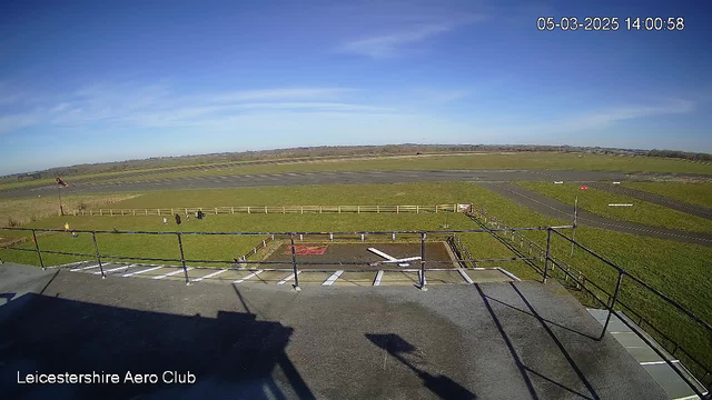 A view from an elevated platform overlooking an expansive green field with a cropped grass surface. In the foreground, there is a black railing along the edge, and several people are seated on the grass, likely enjoying the scenery. The background features a tarmac runway extending into the distance. The sky is clear with a few wispy clouds, indicating a bright and sunny day.