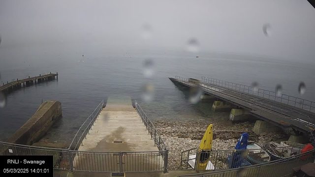 A misty view of a calm body of water, with some raindrops on the camera lens. In the foreground, there are two concrete ramps leading down to the water, one wider and slightly textured, the other narrower, both surrounded by metal railings. To the right, there are several small boats parked, with a yellow kayak and a blue boat visible. The background features a distant, hazy shoreline and several small boats or buoys floating in the water.