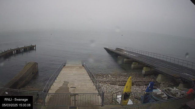 A foggy waterfront scene showing a concrete ramp leading down to the water's edge, flanked by a metal railing. To the left, a wooden pier extends out into the calm sea. Several small boats are visible on the surface of the water, with a few more on the shore, next to a yellow kayak and a blue kayak. The rocky shore is partially visible, and the overall atmosphere is quiet and subdued due to the fog. The date and time in the bottom left corner indicate March 5, 2025, at 15:00:08.