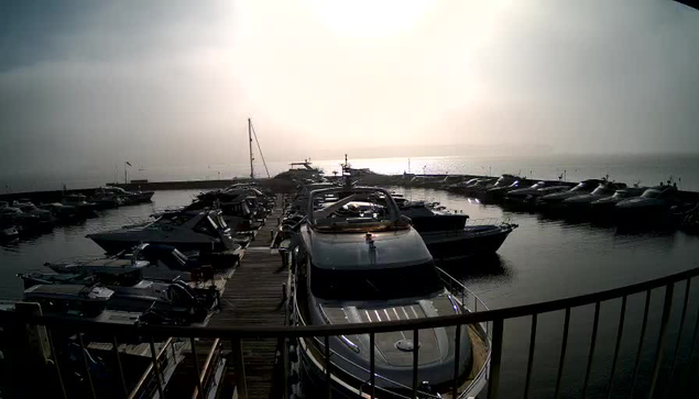 A view of a misty marina at sunrise. Several boats are docked in the calm water, reflecting the light. In the foreground, larger boats are visible, with a wooden dock extending alongside. The background features a hazy sky and water that meets the horizon, creating a serene yet obscured landscape.