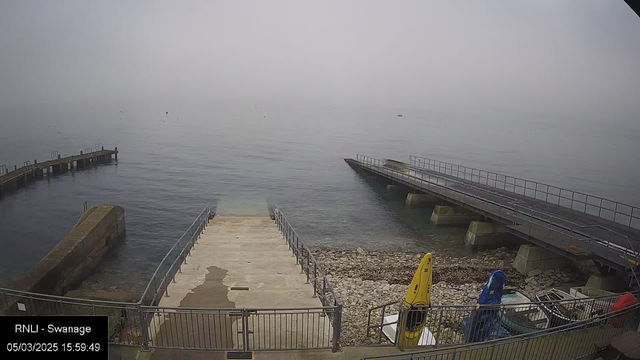 A misty scene at a seaside location with calm water. There are two piers extending into the water on either side. One pier is wooden with a railing, while the other appears to be partially concrete and more robust. In the foreground, a ramp leads down to the water, with steps visible on the left side. The shoreline is rocky, with some colored kayaks (yellow and blue) parked nearby. The background is obscured by fog, making it difficult to see the horizon.