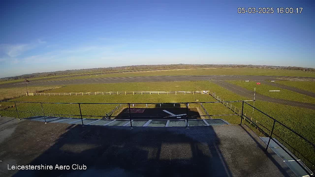 A clear blue sky above a green airfield with a runway marked by gray asphalt and white lines. In the foreground, there is a railing of a viewing platform or building. A small red object is visible near the edge of the runway, and a weather vane stands to the left. The overall scene is well-lit and shows an expansive view of the landscape.