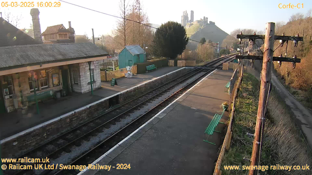 A railway station platform viewed from an elevated angle. The scene features a stone building with a sloped roof on the left, partially shaded by a tree. Green benches line the platform, which has two railway tracks running through it. In the background, the landscape rises to a hill topped with a castle ruin. The sky is clear, and the sunlight casts soft shadows, creating a warm atmosphere. There is a wooden fence and a green shed in the background, along with some vegetation.