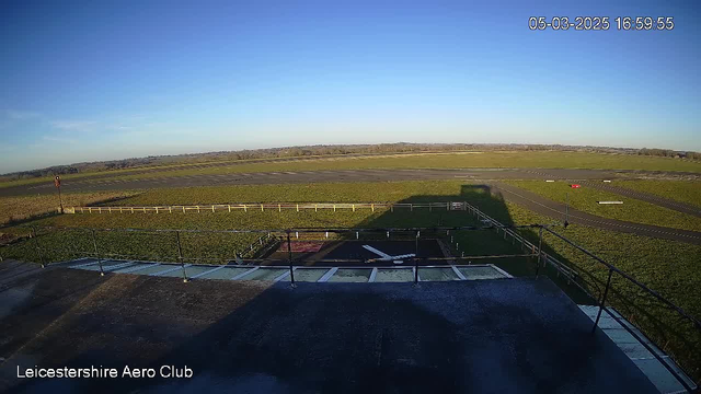 A clear blue sky fills the upper part of the image, transitioning to a green grassy field below. In the foreground, there is a flat rooftop with a railing. The ground features an open grass area that leads to a runway, which is visible in the center. A few white strips are marked on the runway, and a red light is situated on the right side. The scene is bathed in daylight, indicating a sunny, clear atmosphere.