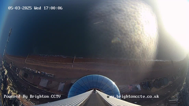 A view from a high vantage point overlooking a beach. The image shows a circular glass structure at the base, with visible support beams extending upwards. Below, the sandy beach stretches alongside a calm body of water. The shoreline is lined with buildings, and some people can be seen walking on the beach. The scene is illuminated by sunlight, creating a shimmering effect on the water's surface. The date and time are displayed in the upper left corner, indicating it is March 5, 2025, at 17:00:06.