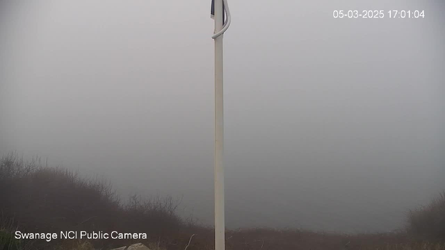 A foggy scene indicating low visibility. On the left, there is a tall white pole with a small attached object. The ground is slightly visible with some dry grass and vegetation. The background is largely obscured by thick fog, making it difficult to discern any distinct features or landforms. The image has a timestamp in the top right corner, reading "05-03-2025 17:01:04," and a watermark in the bottom left corner that says "Swanage NCI Public Camera."