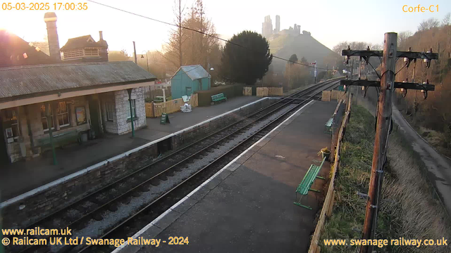 The image shows a railway station platform in the late afternoon. The platform features a stone building with a sloping roof, and wooden benches painted green. A green shed is visible in the background, along with a neatly fenced area. In the distance, a hill rises with the ruins of a castle at its peak, partially shrouded in mist. Power lines run alongside the platform, and the sky is illuminated with a warm golden hue from the setting sun. The scene conveys a peaceful, rural atmosphere.