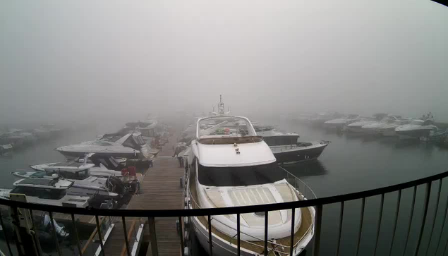 A foggy marina scene with several boats docked. In the foreground, a large white boat with a brown deck is visible, facing away from the viewer. Other boats are scattered in the background, partially obscured by the dense fog. A wooden dock extends into the water, leading to the left side of the image. The water appears calm and reflects the muted light from the foggy surroundings. A railing is present in the foreground, framing the view.