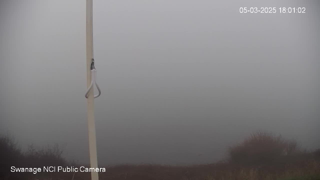 The image shows a hazy and foggy scene, with visibility limited. In the foreground, there is a vertical white pole, which has a hook attached to it. Beneath the pole, the ground is mostly obscured by the fog, making it difficult to discern details. The background merges into a greyish mist, indicating a body of water or a horizon that is not clearly visible. The date and time are displayed in the top right corner.