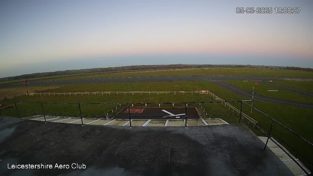 A view from a high vantage point, showing an open field with a small airport runway. In the foreground, there is a railing and a patch of pavement. The runway is marked with lines and has a red light signaling at the far left. The sky displays soft pastel colors, transitioning from light blue to pink as it approaches sunset. The landscape is mostly flat with a few distant trees lining the horizon. The date and time are displayed in the top right corner.