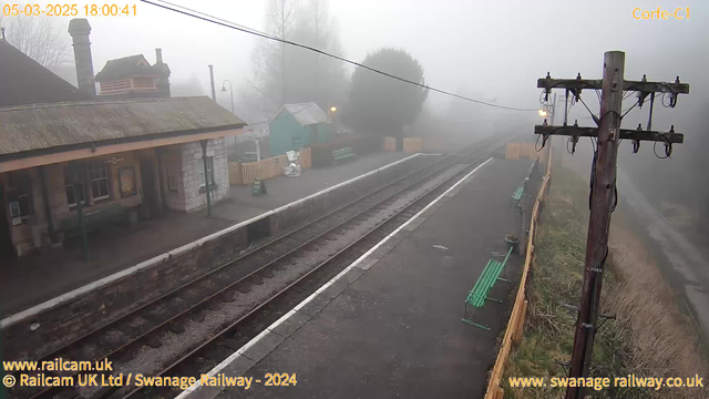 A foggy railway station scene at Corfe Castle. The platform includes several green benches along the edges. A wooden post with electrical lines stands on the right side, and the tracks extend into the mist, leading away from the camera. A small building, likely a ticket office, is visible on the left with a sloped roof and a chimney. The background features trees and fences partially obscured by fog. The overall atmosphere is quiet and somewhat eerie due to the thick mist.