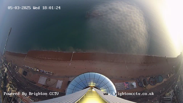 A view looking down from a high vantage point, showing Brighton beach and the sea. The sandy beach is lined with a promenade and various colorful buildings. Some structures, possibly amusement rides, are visible near the water. The coastline curves gently, and there are patches of people on the beach. Above, the sky appears hazy with soft colors, indicating either early morning or late afternoon light.