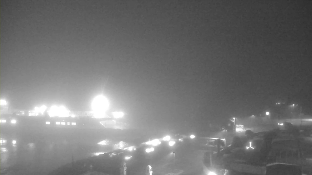 A dark and foggy scene depicting a harbor or pier area. Bright lights from boats and structures create a hazy glow, while the water reflects this light faintly. The foreground shows a dark, indistinct shoreline with silhouettes of boats or structures, partially obscured by fog.