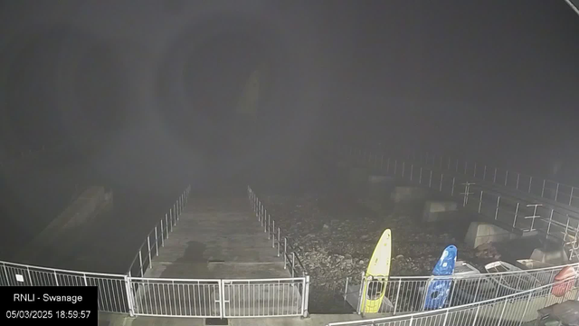 A dimly lit scene captures a wooden pier extending into a body of water, with fog and darkness obscuring visibility. To the left, there are two brightly colored kayaks, one yellow and one blue, positioned on the ground next to the pier. The pier is enclosed with a low fence, and the rocky shoreline is visible alongside it. The atmosphere is quiet and moody, suggesting evening or night time. A timestamp in the lower left corner indicates the date and time as 05/03/2025 at 18:59:57.