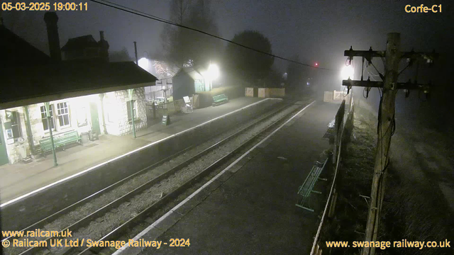 A dimly lit train station scene shrouded in fog. There is a stone building on the left with a green bench in front. The platform has two parallel tracks, and an illuminated signal in the background emits a red light. There are a few more benches along the platform, and a wooden fence is visible in the distance. The atmosphere is quiet and mysterious, with muted colors due to the fog.