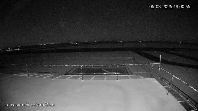 A nighttime view from a webcam showing the Leicestershire Aero Club. The image appears dark, with limited visibility. A flat rooftop edge with a railing is in the foreground, and below is an airstrip or runway area, which is mostly unlit. Far in the distance, faint lights or reflections can be seen, likely from buildings or vehicles. The camera date and time display is in the top right corner, indicating it is March 5, 2025, at 19:00:55.