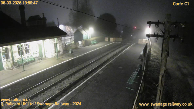 A foggy scene at a train station. The platform is illuminated by lights, revealing a stone structure with a green door and large windows. There are benches along the platform and a sign that reads "Way Out." In the background, a faint red signal light is visible, along with a pole carrying electrical lines. The atmosphere is dreary due to the heavy fog, and proximity outlines of nearby structures and a fence are present. The image captures the tranquility and solitude of the station at twilight.