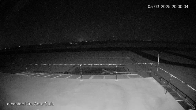 A low-light image taken from a webcam showing a dark outdoor scene. In the foreground, there is a flat surface, possibly a rooftop or platform, with a railing along the edge. The background features a faintly illuminated horizon with scattered city lights in the distance, indicating a nighttime view. The overall tone is dark, with minimal detail visible due to the low lighting conditions. A timestamp indicating the date and time is present in the corner.