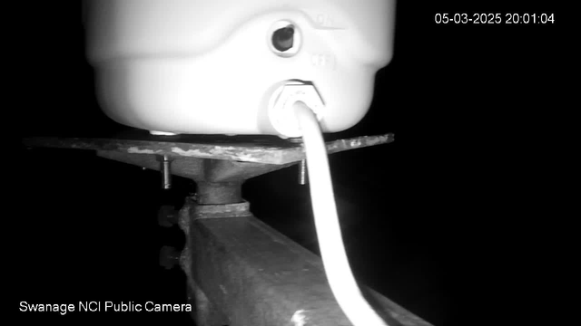 A low-light image showing a white container mounted on a metal base. The container has a visible opening and a connector, with a white cord leading away from it. The background is dark, making the container and the base stand out. Text at the bottom indicates "Swanage NCI Public Camera" and a timestamp at the top right corner reads "05-03-2025 20:01:04."