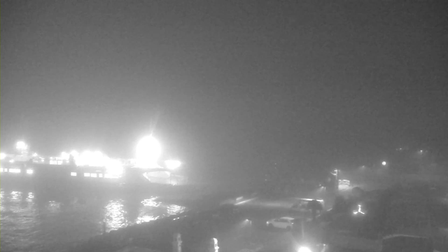 A foggy scene near a body of water, with a faintly illuminated pier on the left. The water reflects soft light, and several vehicles are parked along the shore, barely visible through the fog. The background features indistinct shapes of boats and structures emerging from the mist.