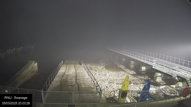 A foggy scene at a waterfront. In the foreground, there is a pathway leading down to the water, bordered by a railing. To the right, two kayaks in bright colors—one yellow and one blue—are visible on a rocky shore. The background shows a partially obscured pier extending out over the water, with a few support pillars visible and dim lighting illuminating the structure. The atmosphere is misty and the visibility is low.