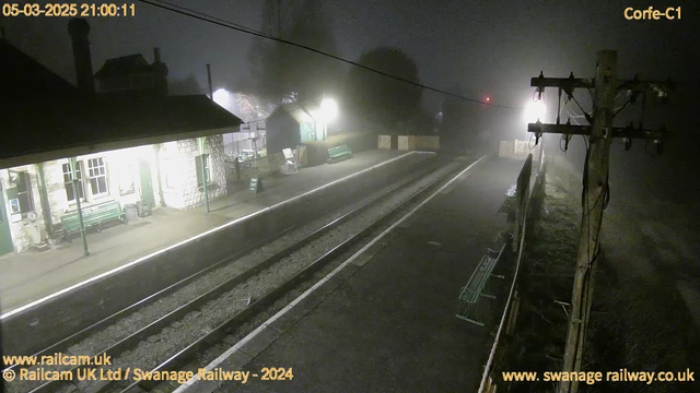 A foggy railway station at night, with dim lighting. The scene includes a stone platform with a green bench, a small building, and a sign that reads "WAY OUT." There are train tracks running through the middle, and faint outlines of trees and a fence are visible in the background. A utility pole with wires stands to the right, and the light from an overhead source creates a soft glow in the fog.