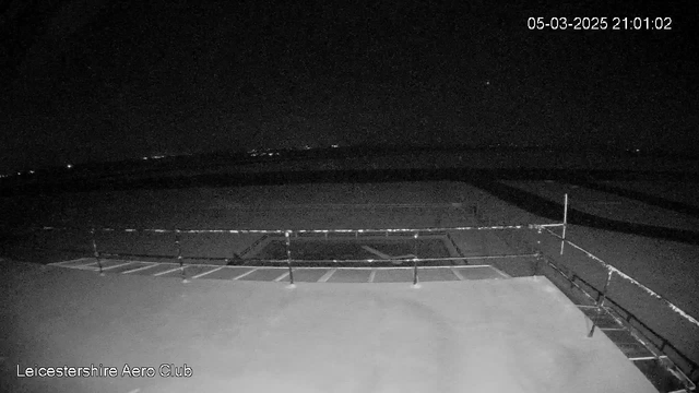 A dark scene captured by a webcam, showing a flat surface that appears to be a runway or tarmac. A railing runs along the front edge, and there are markings on the ground, suggesting a landing area. The image is primarily in shades of black and gray, giving it a nighttime appearance with minimal visibility. In the background, faint lights can be seen in the distance, indicating possibly distant structures or vehicles. A timestamp in the upper right corner shows the date and time as March 5, 2025, at 21:01:02. 