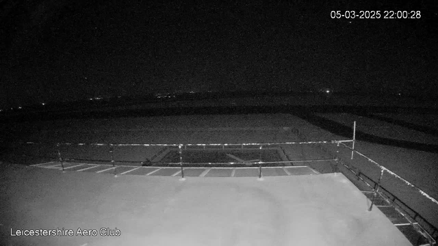 A dark scene captured from a webcam at Leicestershire Aero Club. The image shows a flat surface, likely a runway, with faint outlines of a railing in the foreground. The background is mostly black with a few distant lights scattered across the horizon, suggesting a nighttime view. The date and time are displayed in the top right corner.