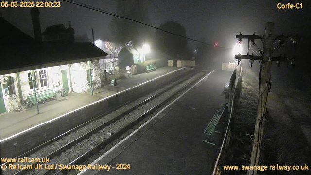 A dimly lit train station platform is visible, shrouded in fog. The left side features a building with large windows and a green bench outside. There is a sign reading "WAY OUT" on the platform. The tracks extend into the distance, while nearby, a wooden fence encloses a small area with another green bench. A utility pole with wires stands to the right. The atmosphere is quiet and eerie, illuminated by soft yellow lights from the building and further down the platform.