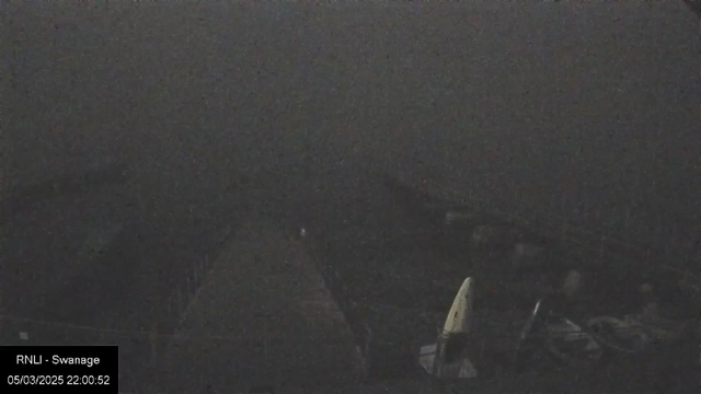 A dark, foggy scene featuring a deserted wooden pier extending into the water. The outlines of boats and objects are faintly visible, creating an ominous atmosphere. There is little visibility, with the background blending into a grayish haze. The date and time in the bottom left corner indicate it is 10:00 PM on March 5, 2025.