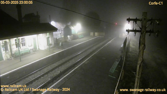 A dimly lit railway station captured at night, shrouded in thick fog. The scene includes a stone building on the left with large windows and a green bench outside. Along the platform, there are railway tracks leading into the mist. A faint light glows from the right, possibly from a signal, and a wooden utility pole stands on the right side of the image with overhead wires. The atmosphere is quiet and eerie, with limited visibility due to the fog.