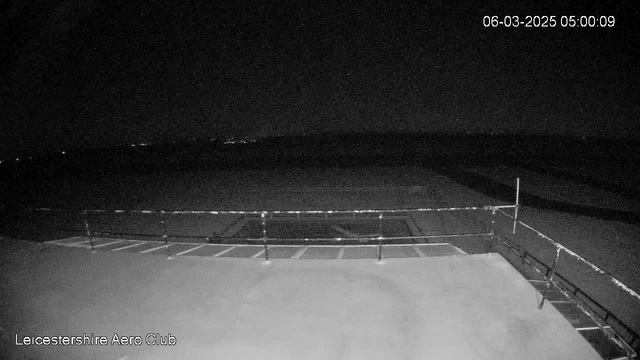 A dark scene captured by a webcam at the Leicestershire Aero Club. The image shows a flat roof area with a railing in the foreground. In the background, there are faint lights indicating a distant horizon, suggesting an open area or possibly a runway. The time is noted as 5:00 AM on March 6, 2025. The overall atmosphere is quiet and empty, with no discernible activity.